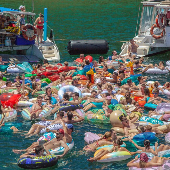 A large group of people relax on colorful inflatable rafts and pool floats in blue water near two docked boats, enjoying a sunny day at a crowded floating party.
