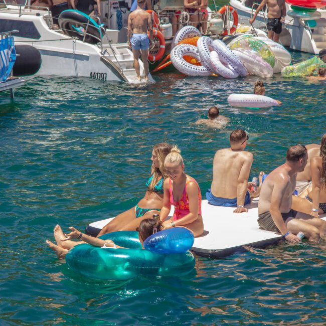 People relax on inflatable floats and a floating dock in clear blue water near boats. Some are sunbathing, chatting, or playing in groups, surrounded by colorful pool floats and summer scenery.