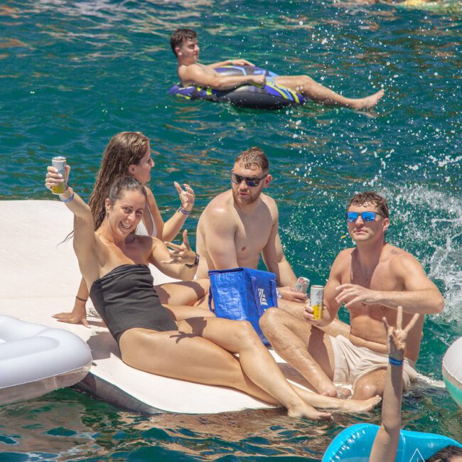 A group of young adults relax on a floating platform in the water, smiling and holding drinks, surrounded by others enjoying inflatables and swimming on a sunny day.