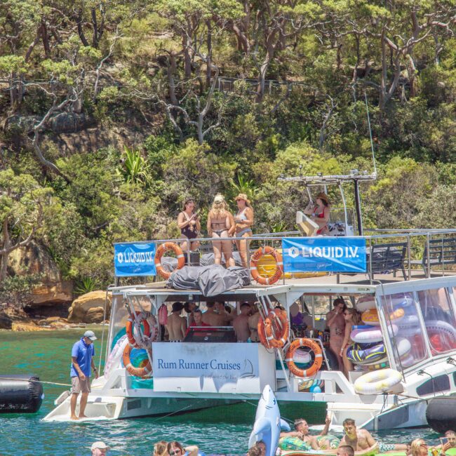 A group of people in swimwear are gathered on a party boat called “Rum Runner Cruises,” anchored near a forested shoreline. Some people are on the upper deck, others are in the water on inflatables. The weather is sunny.