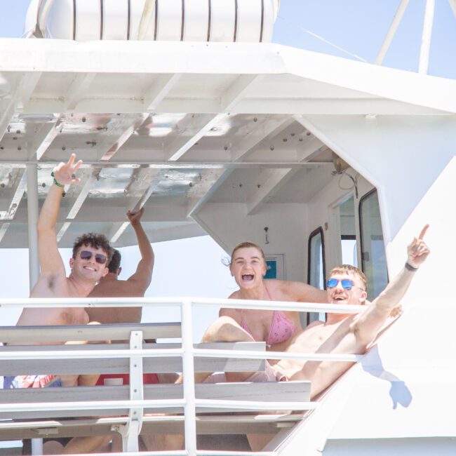 Three people in swimsuits smile and pose on the deck of a white boat, making peace signs and pointing. They appear to be enjoying a sunny day, with others relaxing in the background.