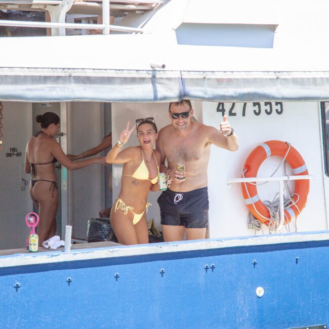 Two people in swimsuits stand on the deck of a boat, smiling and posing with drinks. Other people are visible inside the cabin. A life ring is attached to the blue and white boat. The water is in the background.