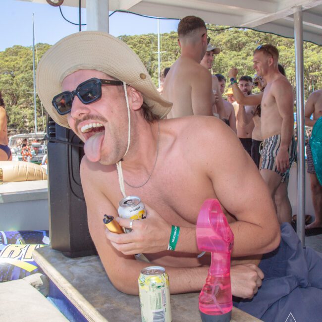 A man in a sunhat and sunglasses leans on a boat railing, sticking out his tongue and holding drinks, while other people in swimwear socialize in the background at a sunny outdoor party.