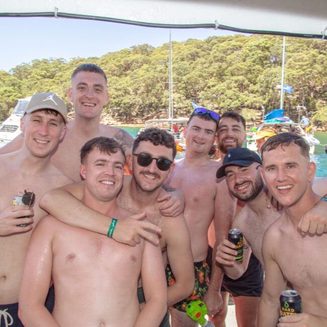 A group of eight smiling young men in swimwear pose together on a boat, holding drinks, with other boats and people enjoying the water and forested shoreline in the background.