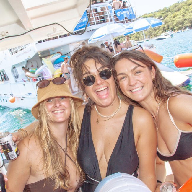 Three smiling women in swimsuits pose together on a boat, holding drinks and a pool float, with other people, another boat, and water activities visible in the sunny background.