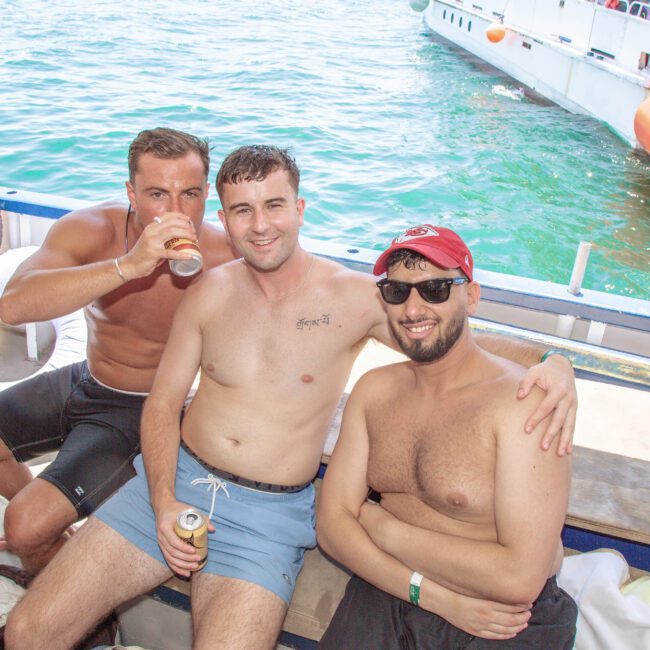 Three young men in swim trunks relax on a boat by the water, smiling at the camera. One drinks from a can, while the others hold cans and look cheerful. Another boat is docked in the background.