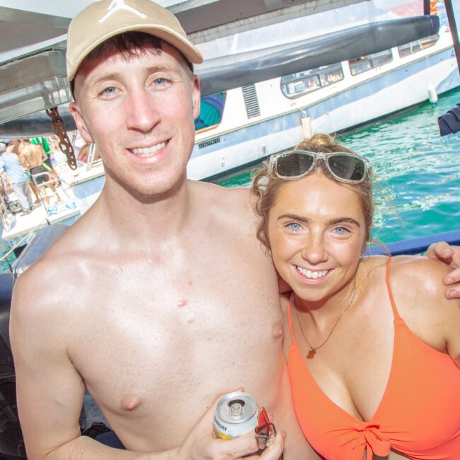 A smiling man and woman in swimwear pose together on a boat, each holding a canned drink. Other people and boats are visible in the background on a sunny day by the water.