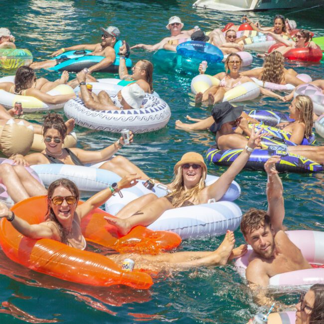 A group of people relax on colorful inflatable pool floats in the water on a sunny day, smiling, waving, and enjoying a lively outdoor gathering.