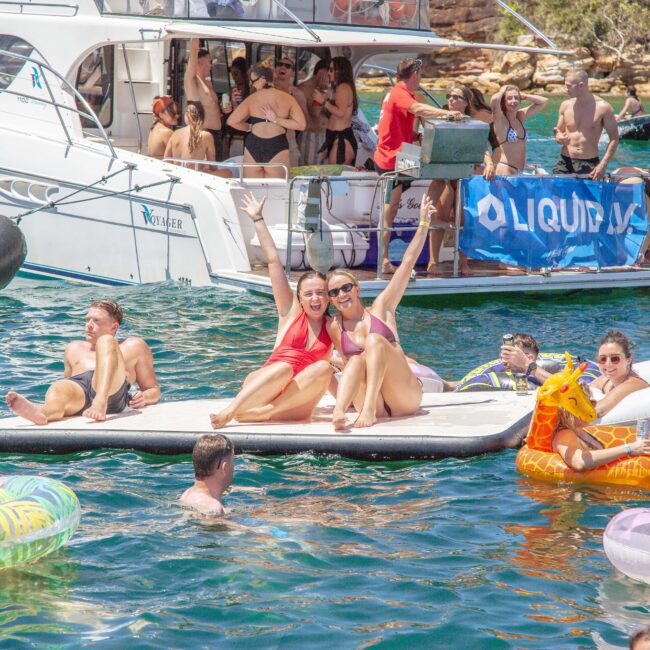 A lively group enjoys a boat party on a sunny day; people relax on inflatables and a floating mat in the water, while others gather and socialize on a yacht in the background.