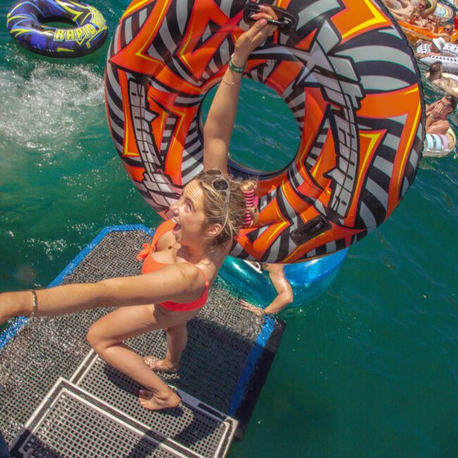 A woman in a red swimsuit smiles and holds up a colorful inflatable tube while standing on a platform by the water, surrounded by people floating on inflatables at a lively outdoor event.