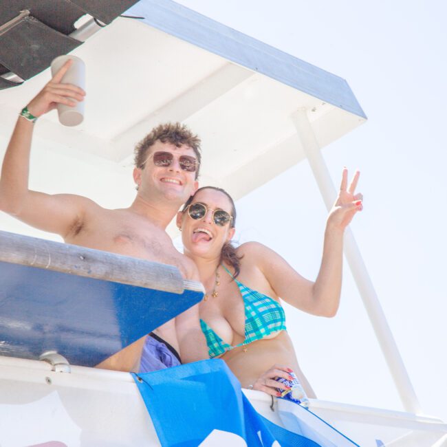 A smiling man and woman in swimsuits pose together on a lifeguard stand, flashing peace signs. The man holds a drink and wears sunglasses, while the woman wears a blue bikini top and sunglasses. It's a bright, sunny day.