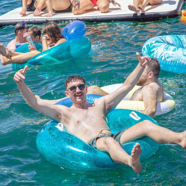 A group of people relax and smile on colorful inflatable floats in a sunny, blue body of water. One man in the foreground cheers with both arms raised, looking joyful and relaxed.