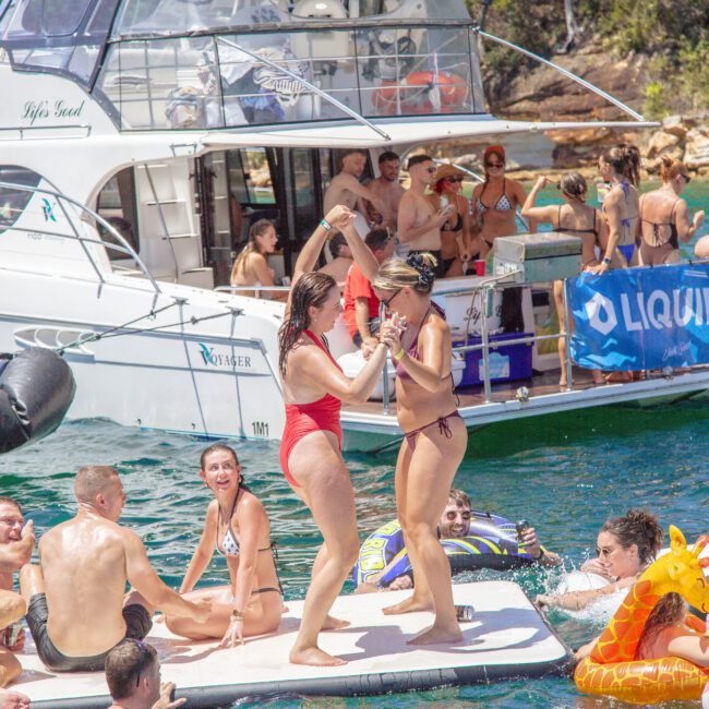 A group of people in swimsuits enjoy a party on a boat and floating platforms on the water, with some dancing, relaxing, and swimming near a forested shoreline under sunny skies.