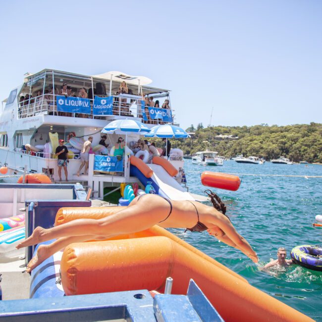 A woman in a bikini dives off an inflatable platform toward the water, while people socialize on a large boat nearby. Several inflatables and smaller boats float on the blue water under a sunny sky.