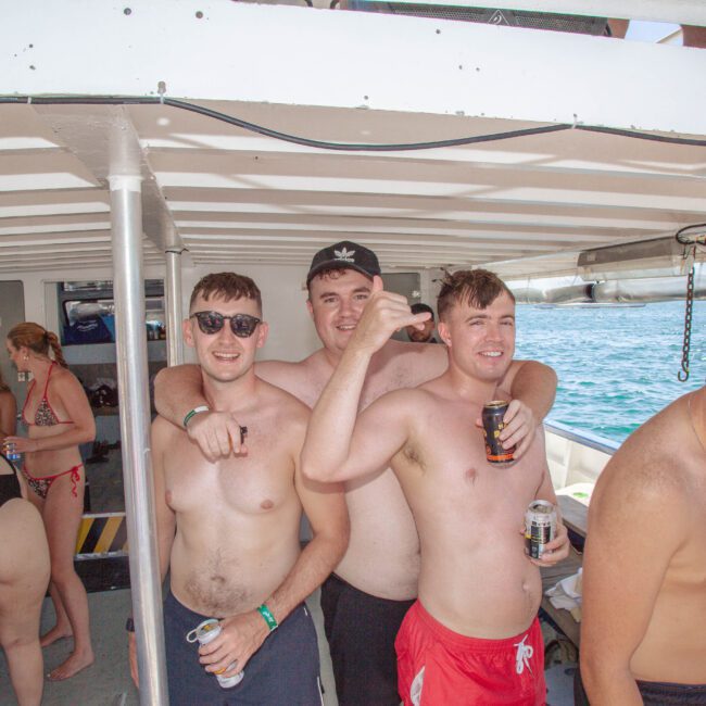A group of young men in swimwear smile and pose with drinks on a boat. Other people in swimsuits are sitting and socializing in the background. The boat is on clear blue water under a sunny sky.