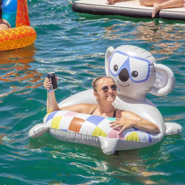A woman in sunglasses smiles and holds up a drink while relaxing on a koala-shaped pool float in the water. Other people on colorful floats and a dock are visible in the background. The water is bright and clear.