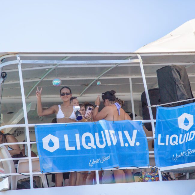 A group of people on the upper deck of a boat decorated with blue "LIQUID I.V." banners; one person in sunglasses makes a peace sign while others socialize in swimwear on a sunny day.