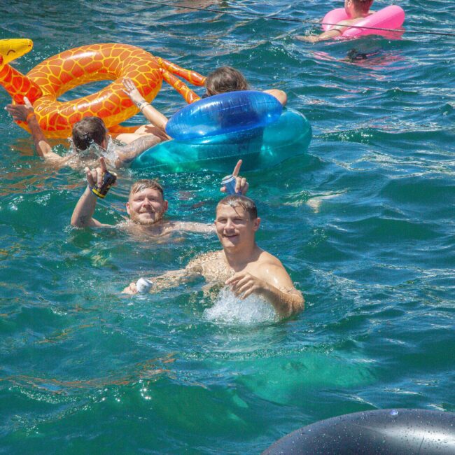 People swimming in blue water with inflatable toys, including a giraffe and a blue ring. Two smiling men in the foreground pose and gesture while others enjoy the water in the background.