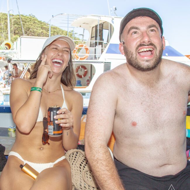 A woman in a white bikini and cap and a shirtless man in a cap laugh and smile while sitting on a boat, with other boats and water in the background on a sunny day.