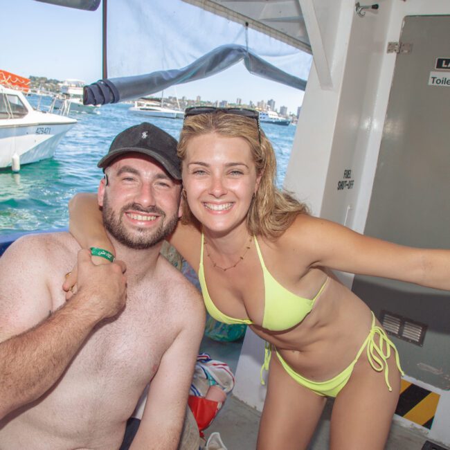 A smiling man and woman in swimwear pose together on a boat, with the woman’s arm around the man’s shoulder. Boats and blue water are visible in the background, and a sign for "Ladies Toilets" is on the wall behind them.