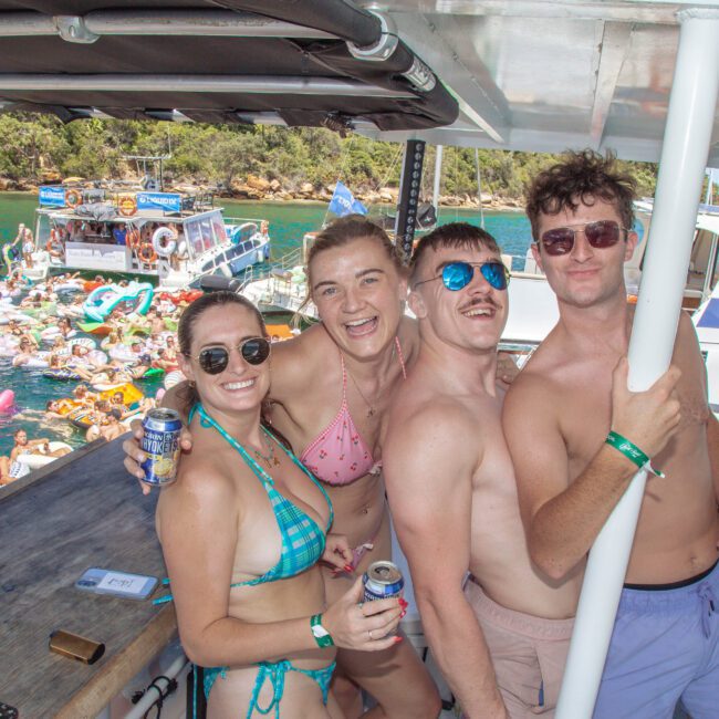 Four smiling people in swimsuits pose together on a boat, holding drinks. Behind them, many people float in the water on inflatables near other boats, enjoying a sunny day. Trees and blue skies can be seen in the background.