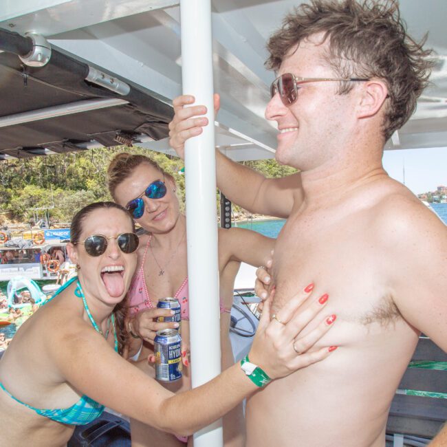 Three people in swimwear are on a boat; two women are smiling and one sticks out her tongue while playfully touching a man’s chest. The background shows water, boats, and people swimming.