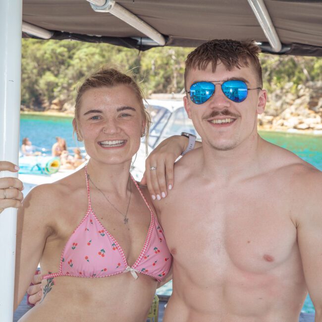 A smiling woman in a pink bikini and a man wearing sunglasses and swim trunks stand together on a boat, with clear blue water and trees visible in the background. The logo “Yacht Social” is in the bottom right corner.