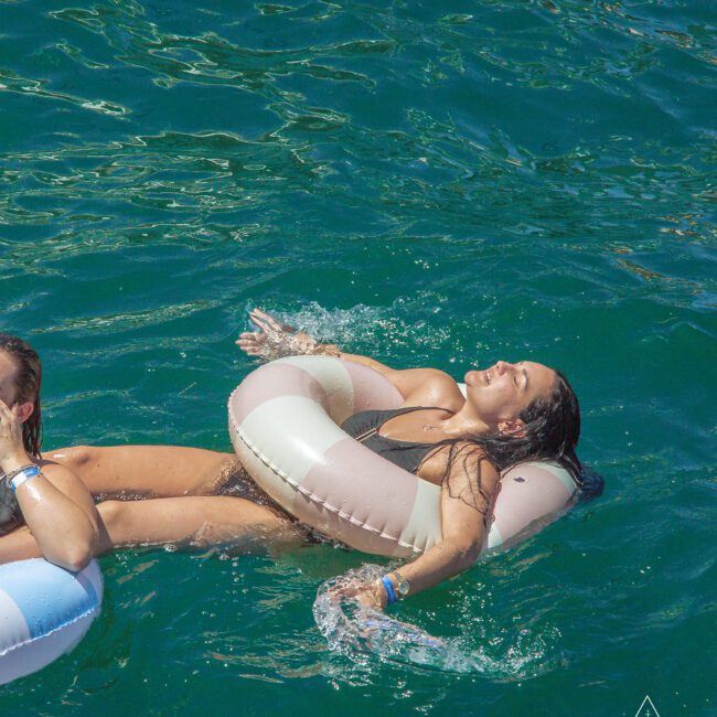A woman in a black swimsuit smiles and relaxes on a striped pool float in the water, enjoying the sunshine. Another person on a float is partially visible beside her. The water is clear and blue.