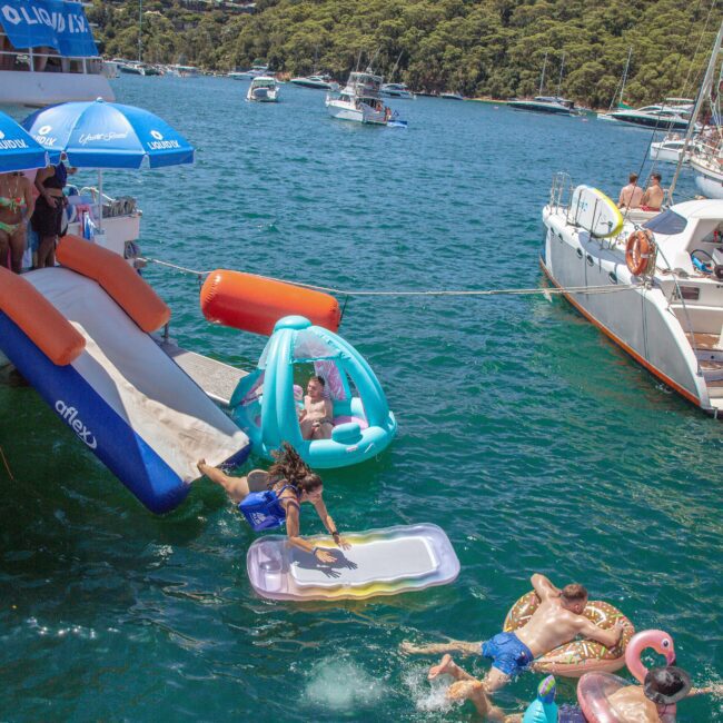 People enjoy a lively boat party on a sunny day, sliding into the water, lounging on inflatables, and socializing near anchored boats surrounded by lush green hills and clear blue water.