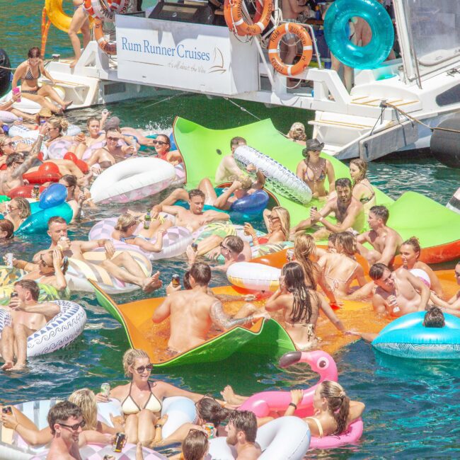 A large group of people relax on colorful floaties and inflatables in the water near a boat labeled "Rum Runner Cruises" on a sunny day, enjoying a lively party atmosphere.