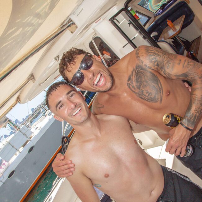 Two smiling, shirtless men pose closely together on a boat. One holds a drink and wears sunglasses; both appear happy. Other passengers and boats are visible in the sunny background.