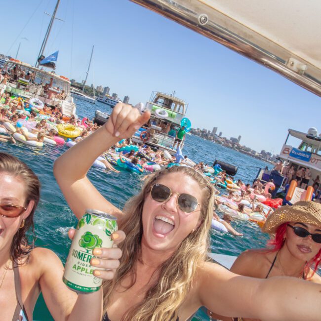 A woman in sunglasses and a black bikini smiles and holds up a drink can on a boat, with other people in swimwear and a crowd of people in the water and on boats in the background on a sunny day.