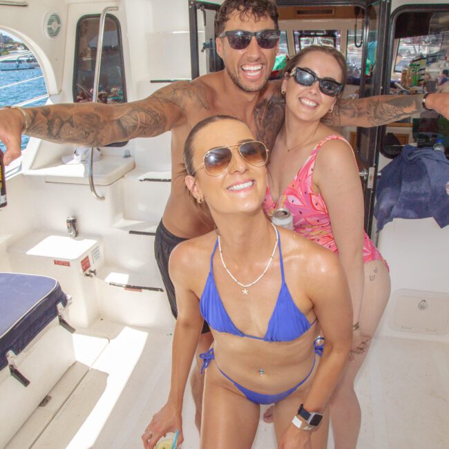 Three smiling people in swimsuits and sunglasses pose on a boat, holding drinks and enjoying a sunny day by the water. A marina and shoreline are visible in the background.