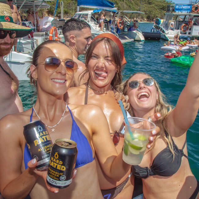 A group of young adults in swimwear smile and pose with drinks on a sunny boat party, surrounded by people, boats, and inflatables on blue water.