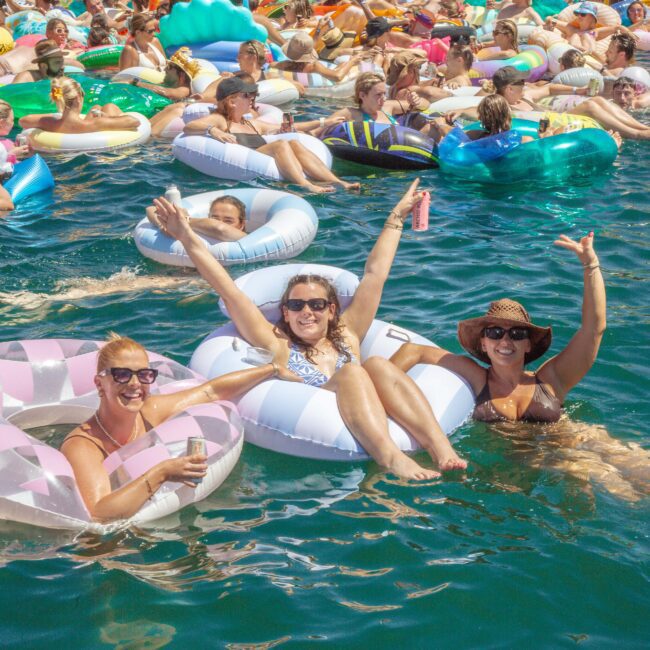 A large group of people float on colorful inflatable tubes in bright blue water, enjoying a sunny day. Three women in the foreground smile and raise their arms, celebrating with drinks. The scene is lively and festive.