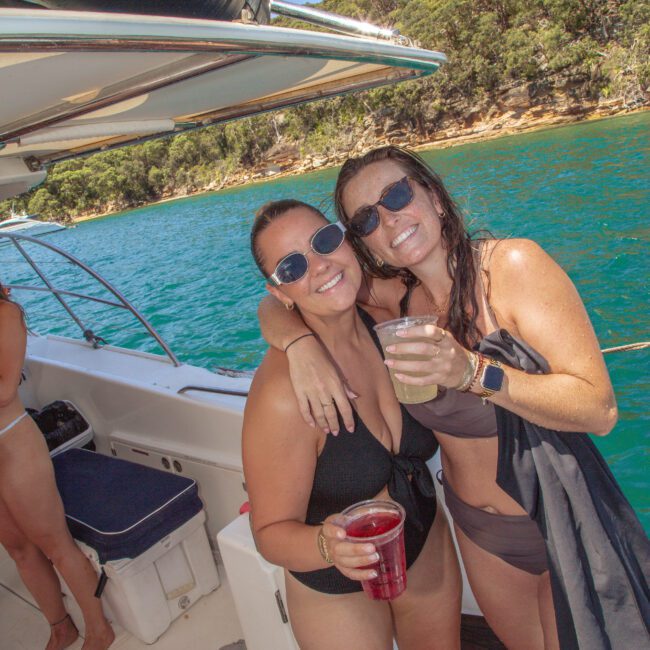 Two women in swimsuits and sunglasses smile and pose with drinks on a boat, with blue water and green trees in the background. Other people and boat details are partially visible. The atmosphere is relaxed and sunny.