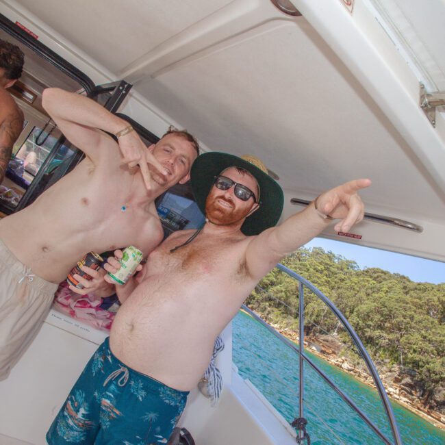 Two men in swim trunks smile and pose on a boat, one pointing out to the water. Both hold drinks, with sun hats and sunglasses. Trees and clear blue water are visible in the background on a sunny day.