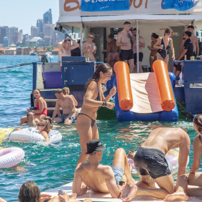 A group of people in swimwear enjoy a summer party on a boat and in the water, surrounded by inflatable pool floats, with a city skyline visible in the background.