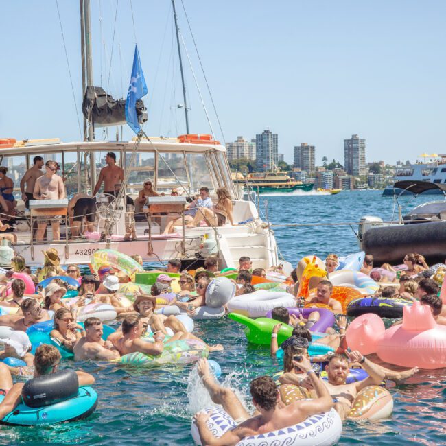 A large group of people enjoying a sunny day on the water, lounging on colorful inflatable floats near boats. A catamaran with more people on deck is anchored nearby, with buildings visible in the background.