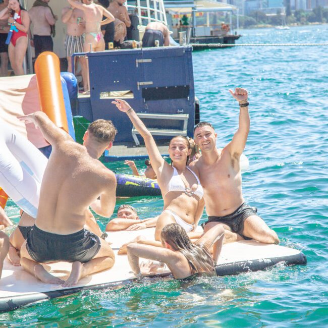 A group of young adults in swimwear relax and have fun on a floating mat in the water, with several people cheering and smiling near a docked boat on a sunny day.