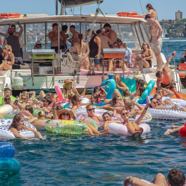 A large group of people relax on colorful inflatable rafts in the water near a boat, enjoying a sunny day. The boat is crowded with people, and the city skyline is visible in the background.