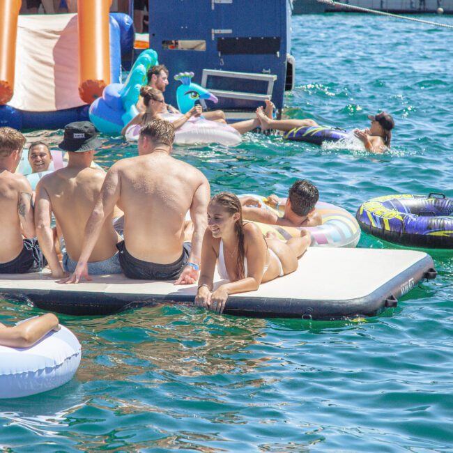 A group of people in swimsuits relax on inflatable rafts and floats in clear blue water near a boat, enjoying a sunny day. Some sit on a floating platform, while others swim or lounge on colorful floaties.
