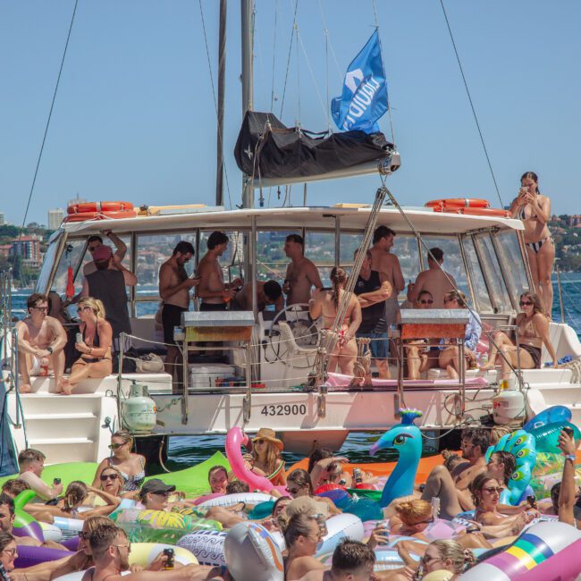 A lively group of people enjoys a party on a catamaran and in the water, surrounded by colorful inflatables, with a sunny city skyline in the background.