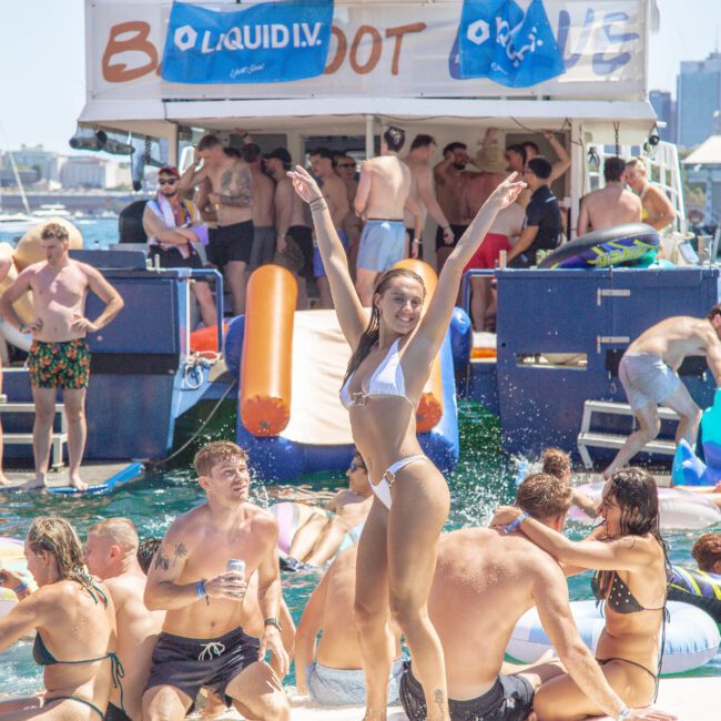 A woman in a white bikini poses with arms raised on a crowded boat party; people swim, relax on inflatables, and mingle in the water, with a city skyline in the background.