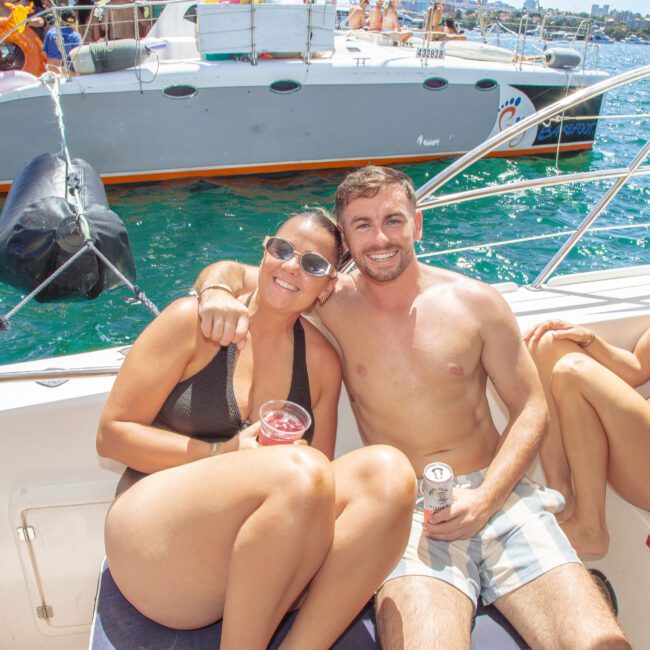 A group of people in swimwear relax and smile on a yacht in sunny weather, holding drinks. Other people are visible on a nearby yacht in the background on the blue water.