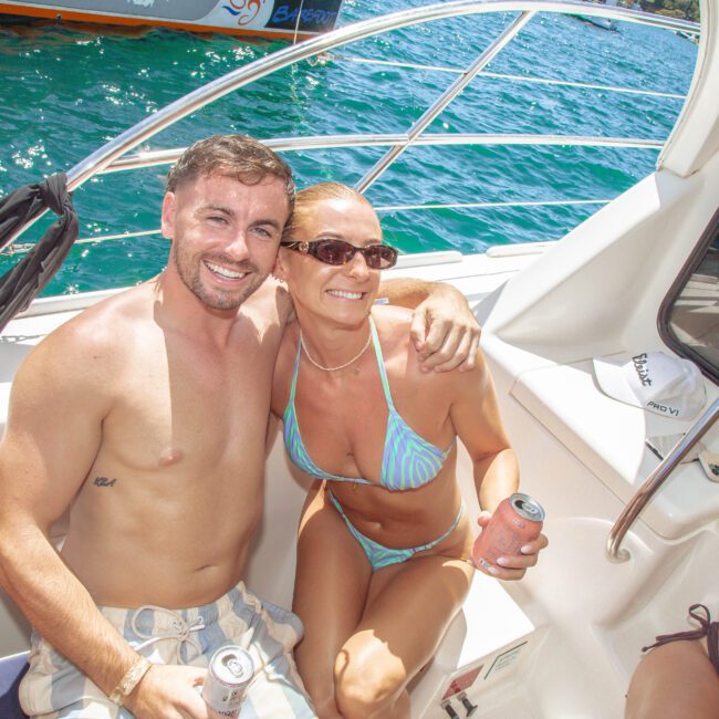 A smiling man and woman in swimwear sit closely together on a boat, holding drinks. The sun is shining, and blue water is visible in the background. Other people are partially visible nearby.
