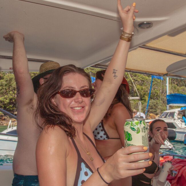 A woman in sunglasses and a polka dot bikini holds a drink and raises her arm while smiling on a boat with friends. Other people in swimsuits are celebrating, and boats and water are visible in the background.