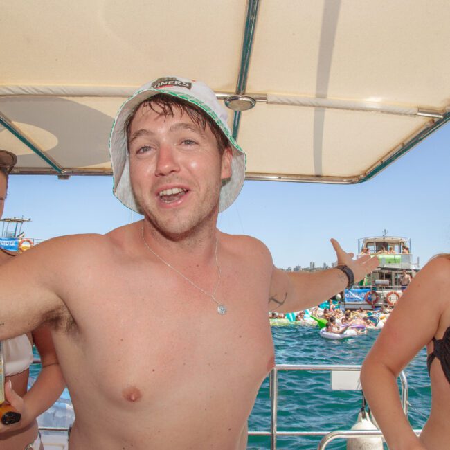 A smiling man in a bucket hat with arms outstretched stands on a boat with two women in swimsuits, with other people, boats, and a sunny blue sky visible in the background.