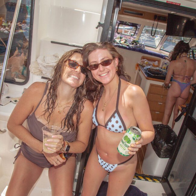 Two women in swimsuits smiling and posing on a boat, holding drinks. Other people are in the background enjoying the sunny day, with water visible outside the boat. The atmosphere is relaxed and festive.