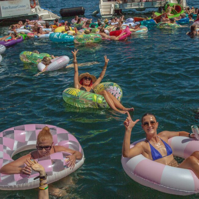 A group of people relaxing on colorful inflatable floats in a crowded lake, smiling and making peace signs. Boats are anchored nearby, and it's a sunny, lively, party-like atmosphere.